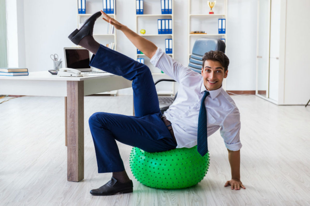 hommeau-bureau-assis sur un ballon-de-exercice-levant-la-jambe-AdobeStock_176789885